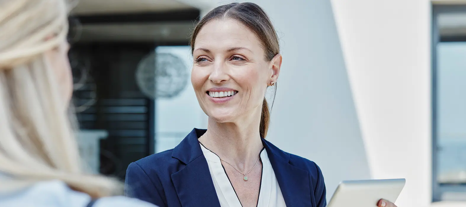 Frau in Businesskleidung hält ein Tablet und spricht mit einer anderen Person im Freien.