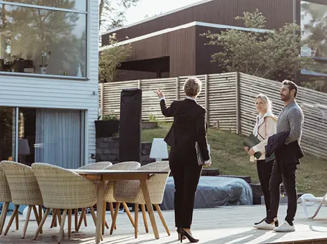 Eine Frau im Anzug zeigt zwei Personen ein modernes Haus mit Terrasse und Gartenmöbeln.